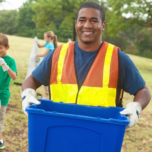 Man in safety vest holding a blue recycling bin outdoors.