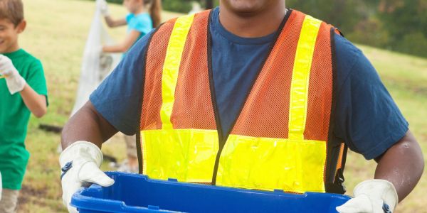 Person wearing safety vest holding a recycling bin outside.