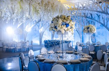Elegant blue and white banquet setup with floral centerpieces and hanging white flowers.