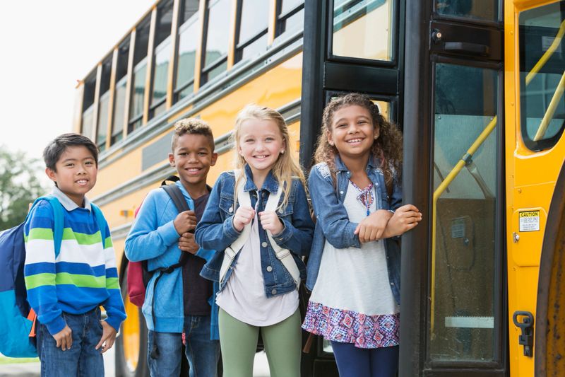 A group of four multi-ethnic school children standing outside the open door of a school bus, smiling at the camera. The mixed race black and Caucasian girl on the right and Asian boy on the left are 8 years old and their friends are 9. They are elementary school students.