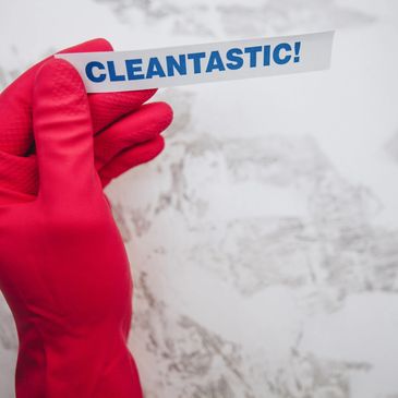 Hand in red glove holds a paper strip with 'CLEANTASTIC!' against a dirty background.
