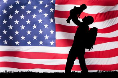 Silhouette of soldier raising his child into the air in front of an American Flag.