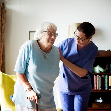 Caregiver assisting elderly woman with walking inside a home.