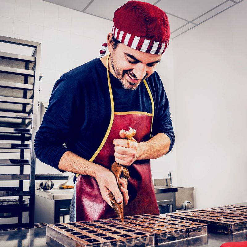 Handmade chocolate pieces. The confectioner, middle age adult man, filling pralines shells in the mold with a cream. Pralines are made from the finest Belgian dark chocolate.