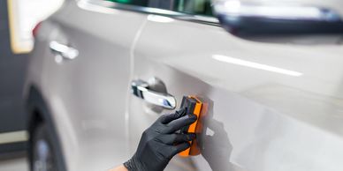 Person wearing black gloves polishing a silver car door with an orange tool.