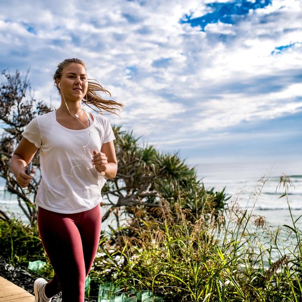 A woman jogging outdoors with earphones, surrounded by greenery and a cloudy sky.