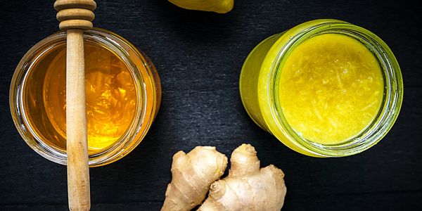 Honey jar with dipper, lemon, fresh ginger, and a jar of ginger paste on a black surface.