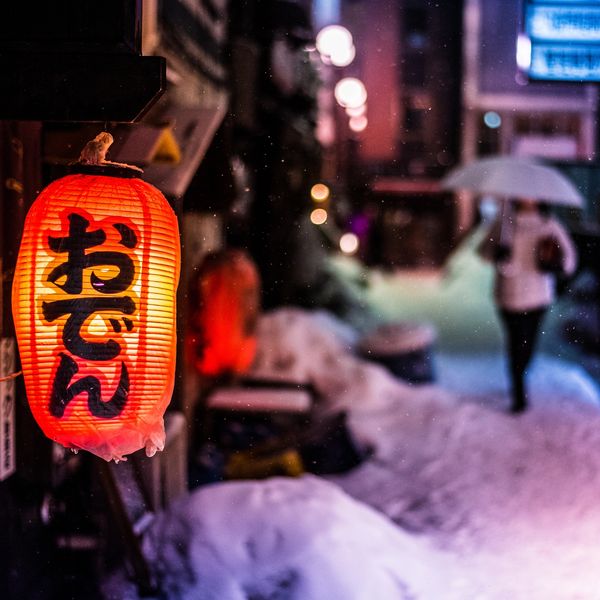 Glowing red Japanese lantern on a snowy street at night with a person holding an umbrella.