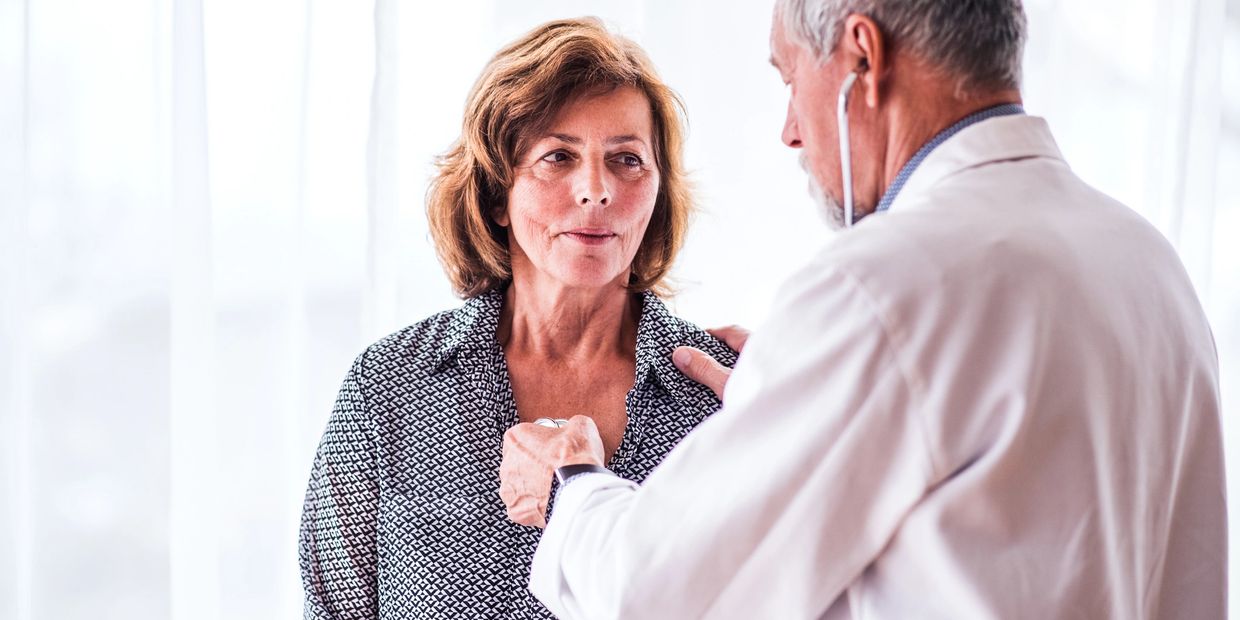 A senior doctor performing a physical exam on a woman, listening to her heart with a stethoscope.