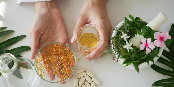 Hands holding capsules and a glass of liquid, surrounded by white pills and flowers.