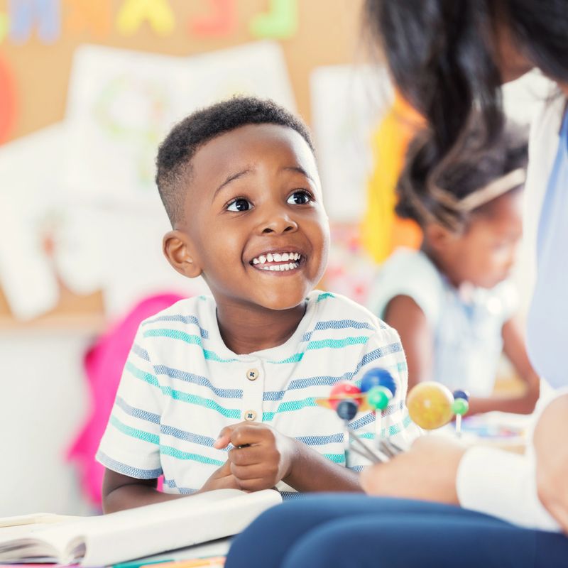 An excited little boy sits at a table in his preschool classroom with his unrecognizable teacher.  He looks up and smiles as he listens to her tell him about a model of the solar system.