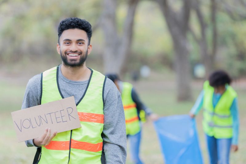 Handsome young male volunteer holds a cardboard 'volunteer' sign while working with his neighbors to cleanup their community park. He is smiling at the camera.