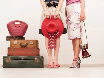 Two women standing beside stacked vintage suitcases with a red hat and old camera.