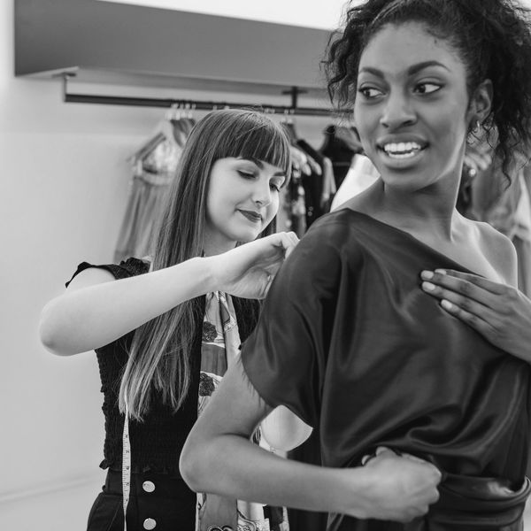 A seamstress fitting a dress on a smiling woman in a boutique.