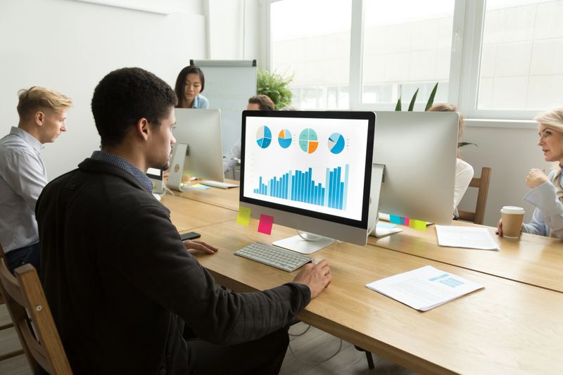 African american project manager working with statistics on computer in multiracial office, black employee analyzing charts graphs report using pc sharing coworking desk with diverse corporate staff