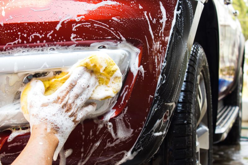 Asian old man using yellow sponge washing front fog light of big new red car. Cleaning background with copy space.
