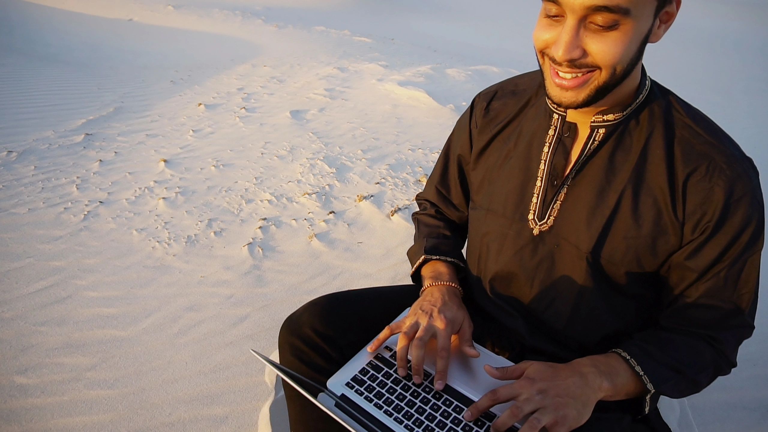 Educated Arab student or young scientist prints on laptop keyboard and working on article, enjoying tranquility and silence of sandy desert sitting on warm sand in open air on summer evening. Swarthy Muslim with short dark hair and beard dressed in black kandur, long spacious dress made of dark cotton with knitted lace cap of hafia, on top of which headscarf and dark brown shoes are tied. Concept of Arab and Muslim men, education and modern technology, business and entrepreneurial activities, Emirati national clothing, good mood and enjoyment of life.