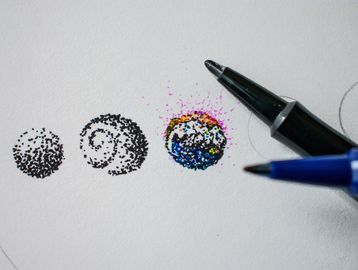 Ink pens creating intricate dotted circle patterns on paper.