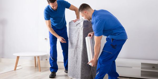 Two movers wrapping furniture in protective plastic inside a modern room.