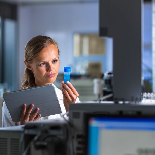 Scientist examining a sample vial while holding a tablet in a laboratory.