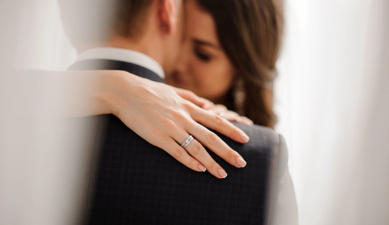 Couple posing outdoors, woman in white dress showing engagement ring.