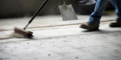 Worker sweeping dust off a concrete floor with a broom and shovel nearby.