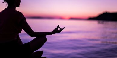 Person meditating by the water at sunset with a serene purple sky.