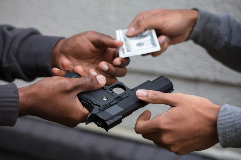Close-up Of Two Hands Exchanging Black Handgun For Banknote