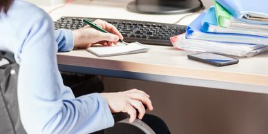 Person in a wheelchair writing notes at a computer desk with files and phone nearby.