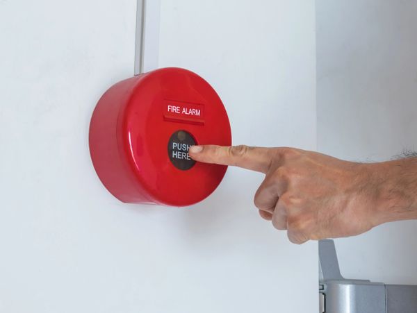 Finger pressing a red fire alarm button on a white wall.