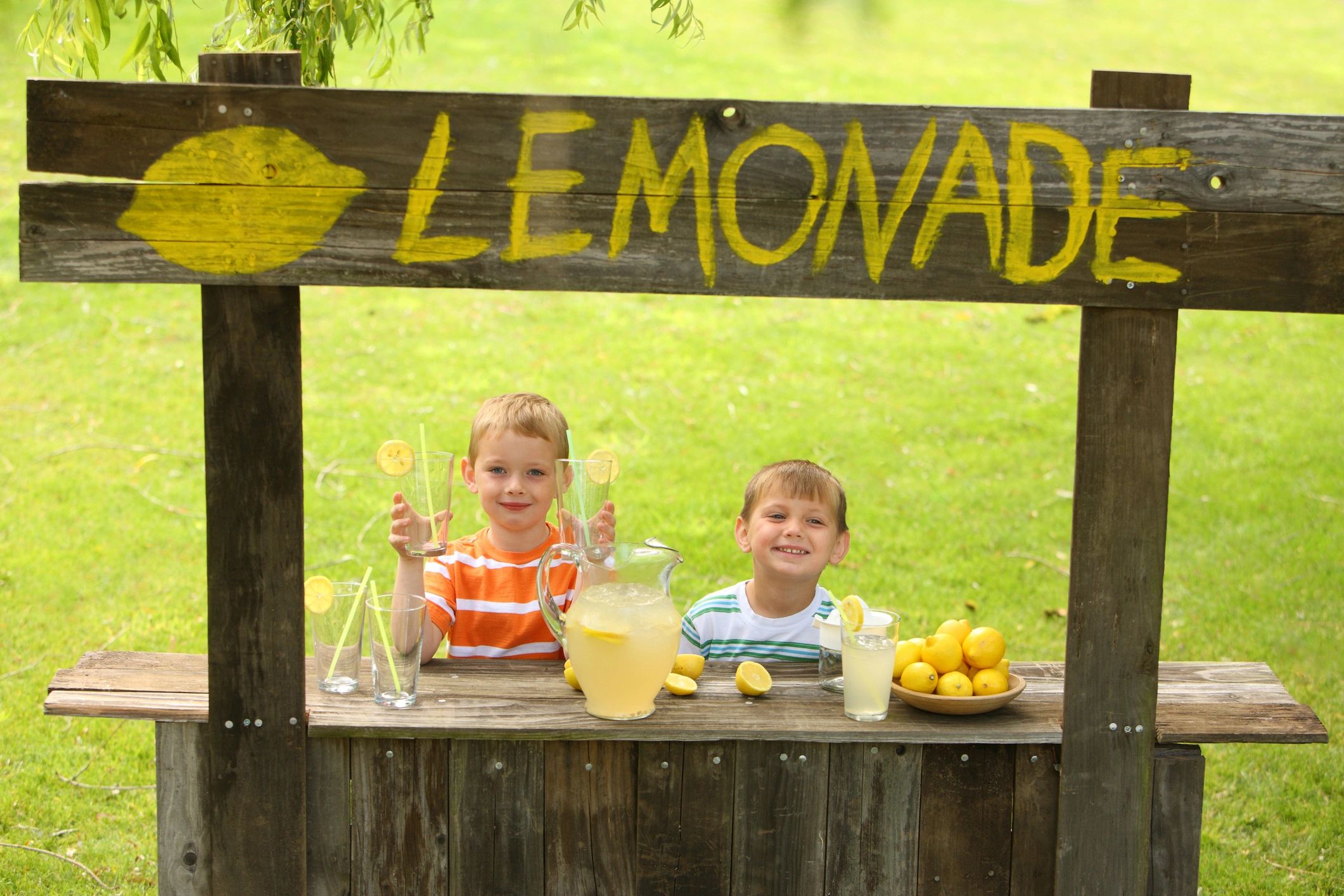 Two kids selling lemonade at a rustic stand outdoors.