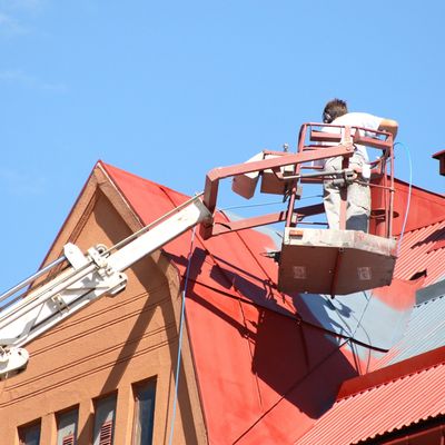 Worker painting a red roof using a hydraulic lift under clear blue sky.