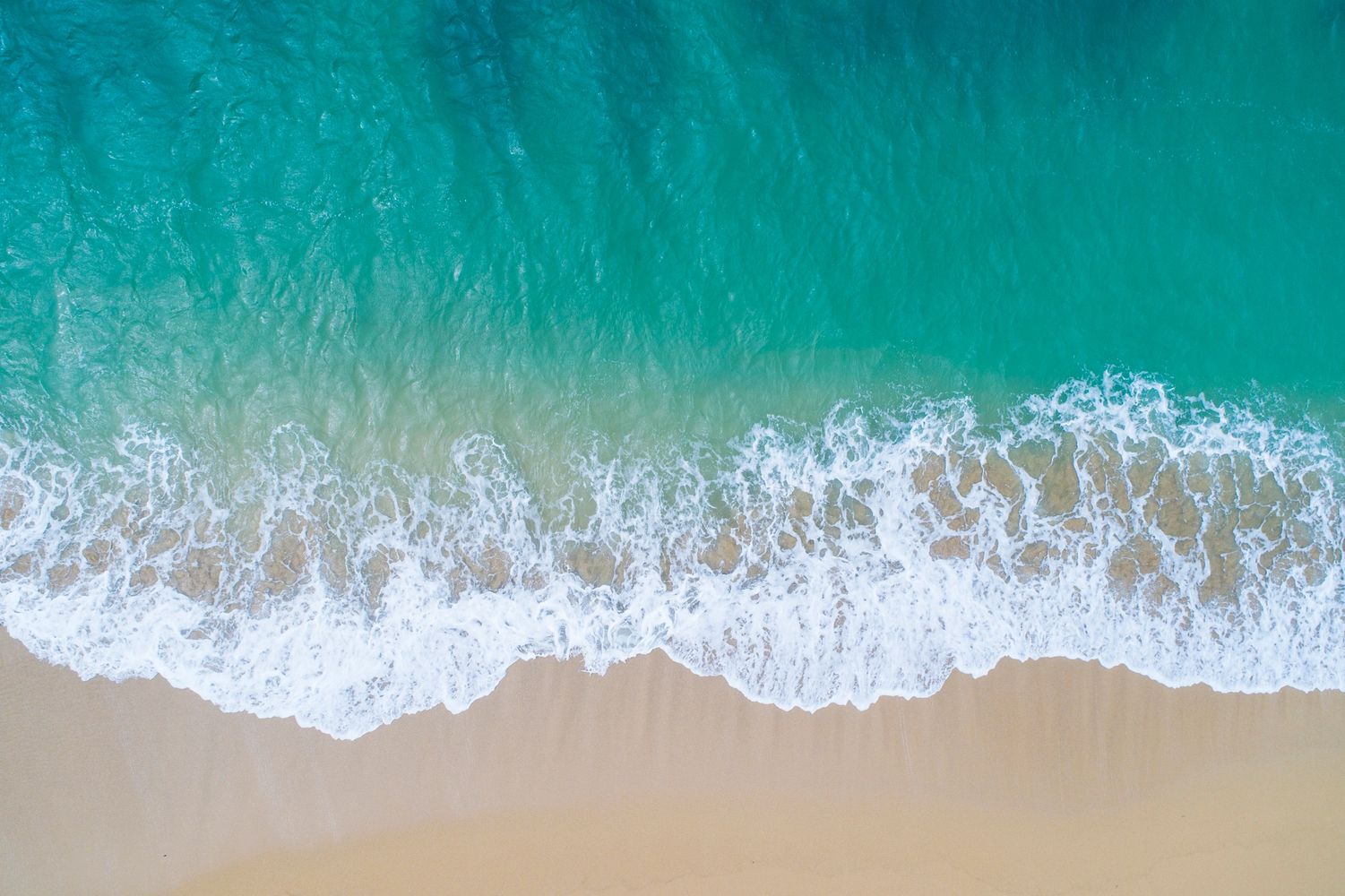 Aerial view of turquoise ocean waves gently crashing on a sandy beach.
