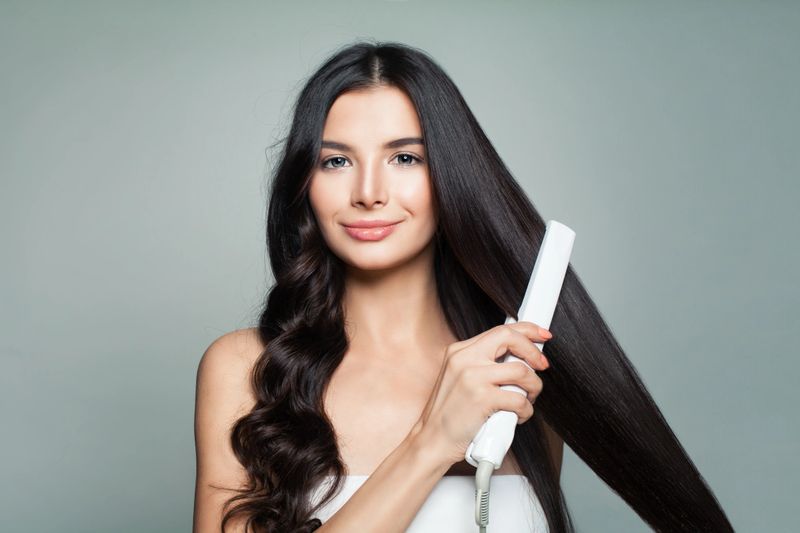 Attractive Woman with Curly Hair and Long Straight Hair Using Hair Straightener. Cute Smiling Girl Straightening Healthy Brunette Hair with Flat Iron. Hair Problem and Haircare Concept