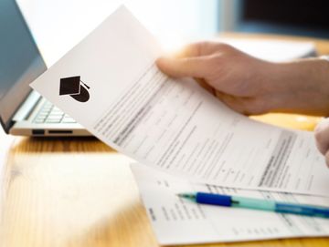 Person reviewing a graduation certificate near a laptop on a wooden desk.