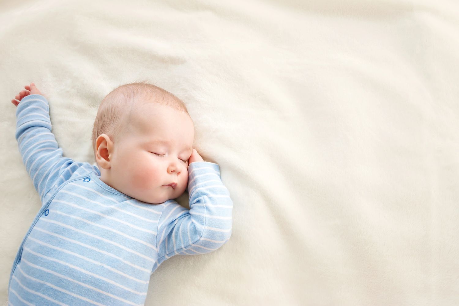 Sleeping baby in a blue striped onesie on a soft white blanket.