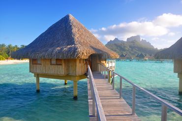 Overwater bungalow with thatched roof in a tropical lagoon under clear blue skies.