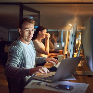 Man working late on laptop in office with colleague in background.
