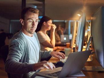 Man working late on a laptop in an office with a woman in the background.