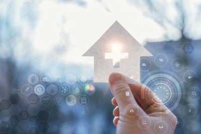Hand holding a house-shaped cutout with a cross symbol, surrounded by healthcare icons.
