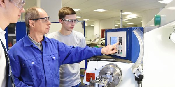 Two men operating a machine in a workshop, wearing safety glasses.