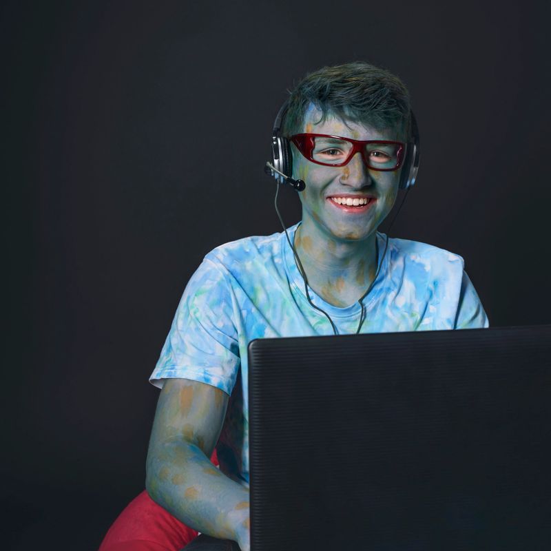 smiling young guy painted with colored paints in glasses, working at the computer