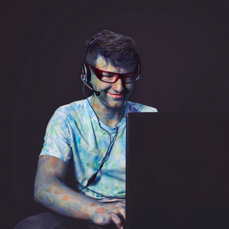 smiling young guy painted with colored paints in glasses, working at the computer