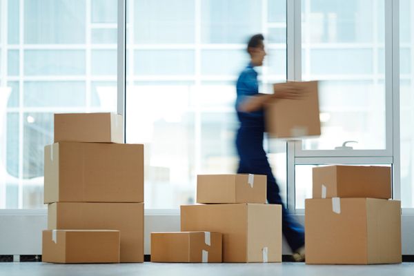 Blurred person carrying a box among stacked cardboard boxes indoors.