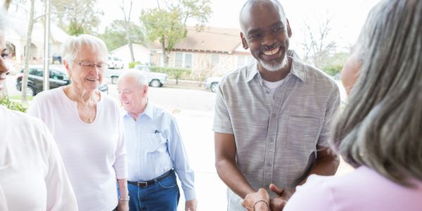 A smiling man warmly greets an older woman while others wait nearby.