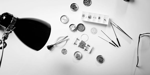 Watchmaker's tools and watch parts spread on a white table under a desk lamp.