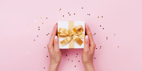 Hands holding a white gift box with a gold ribbon on a pink background with star confetti.