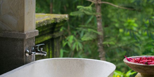 Outdoor stone bathtub with a nature backdrop and flower petals on a nearby stone pedestal.