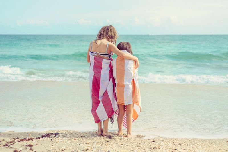 Two children with colorful towels wrapped around them stand at the waters edge of the beach in paradise, beautiful sunny coastal area. Brightly colored towels and affectionate friends or sisters