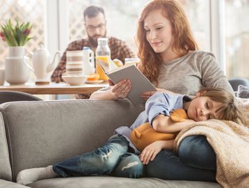 A woman reads a book while a child sleeps on her lap holding a stuffed toy.